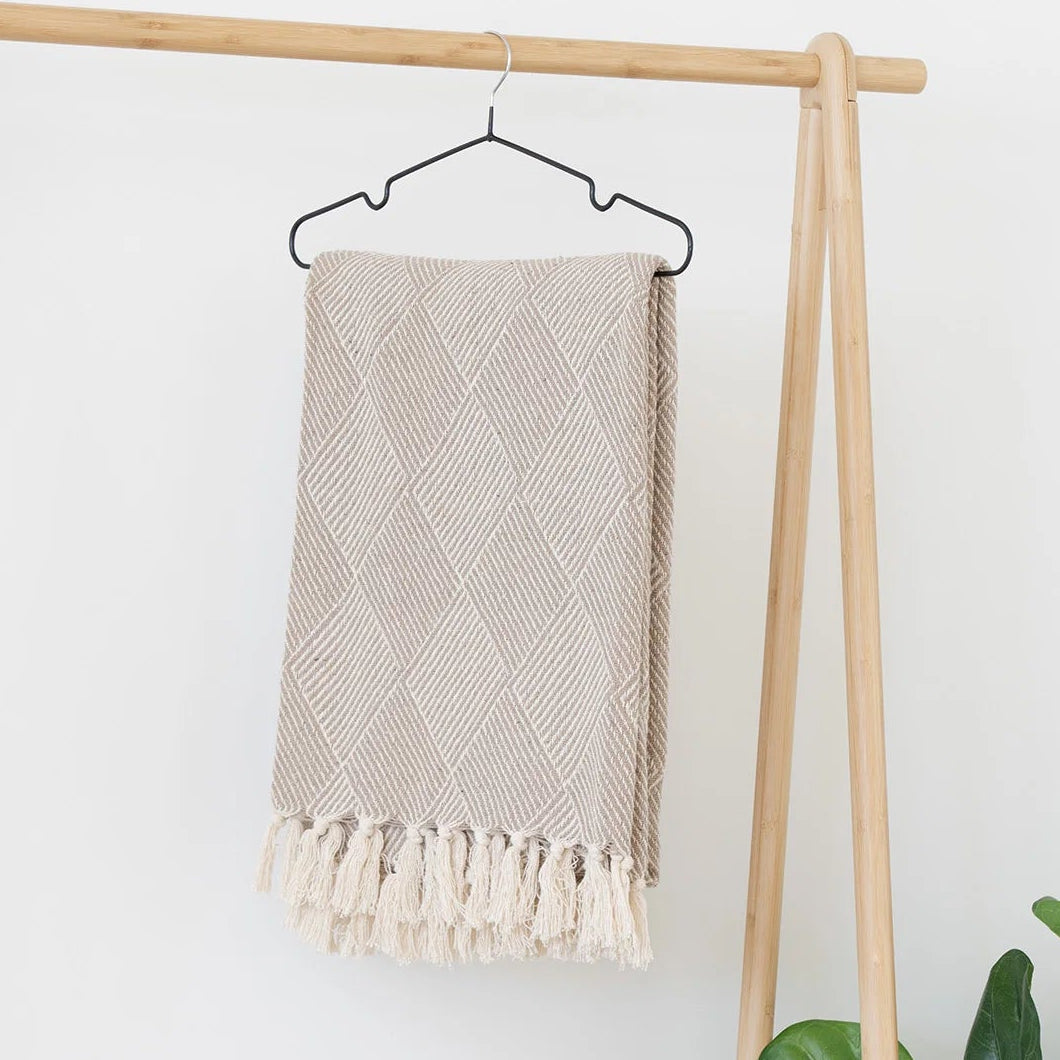 Beige towel with tassels hanging on a wooden rack against a white wall with a plant in the corner.