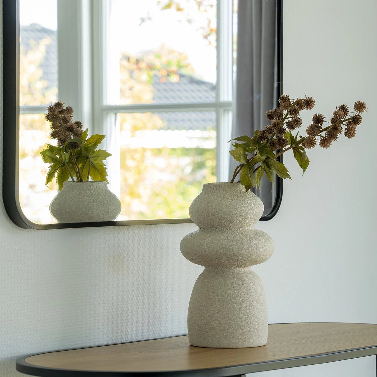 Rectangular mirror with black frame on a wall above a wooden console table with a white vase and dried plants.